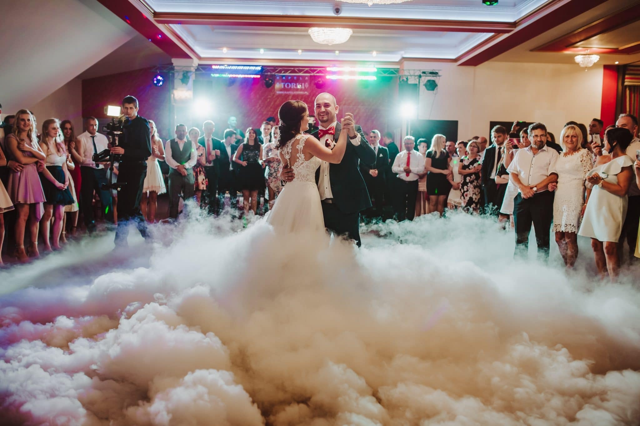 Pierwszy taniec pary młodej w chmurach dymu na sali weselnej, goście obserwują.
