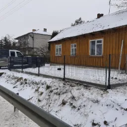 Nowo zamontowane, czarne ogrodzenie panelowe na betonowej podmur&oacute;wce, oddzielające posesję z drewnianym domem od drogi w zimowym krajobrazie.