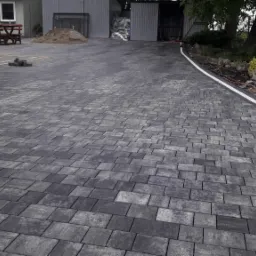 Ułożona szara kostka brukowa na podjeździe, widoczna zagęszczarka wibracyjna oraz obrzeża z kostki granitowej oddzielające nawierzchnię od trawnika.