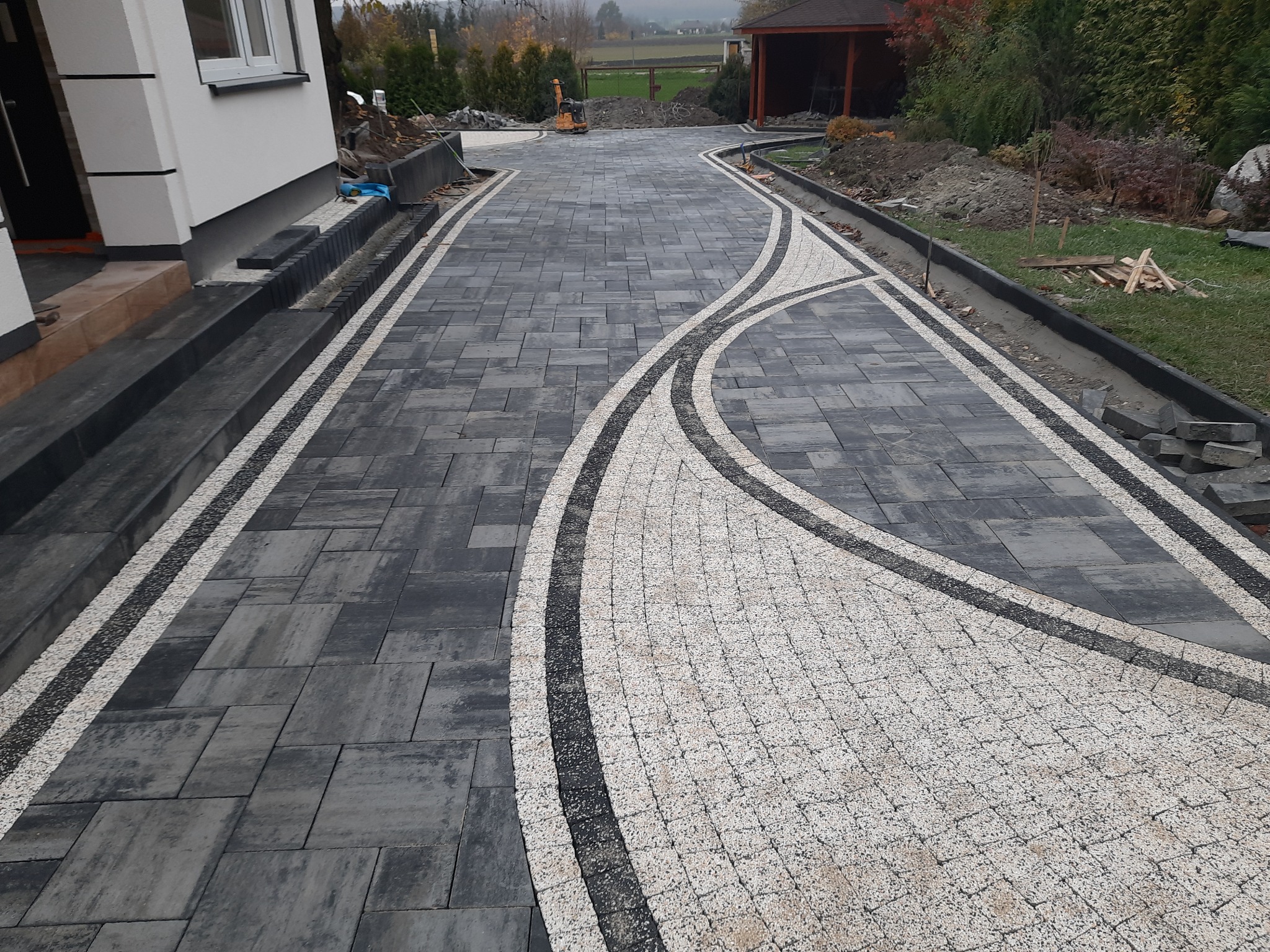 Ułożony podjazd z ciemnoszarej kostki brukowej z dekoracyjnym wzorem z jasnej kostki, obrzeża z kostki, widok na dom i ogród w tle.