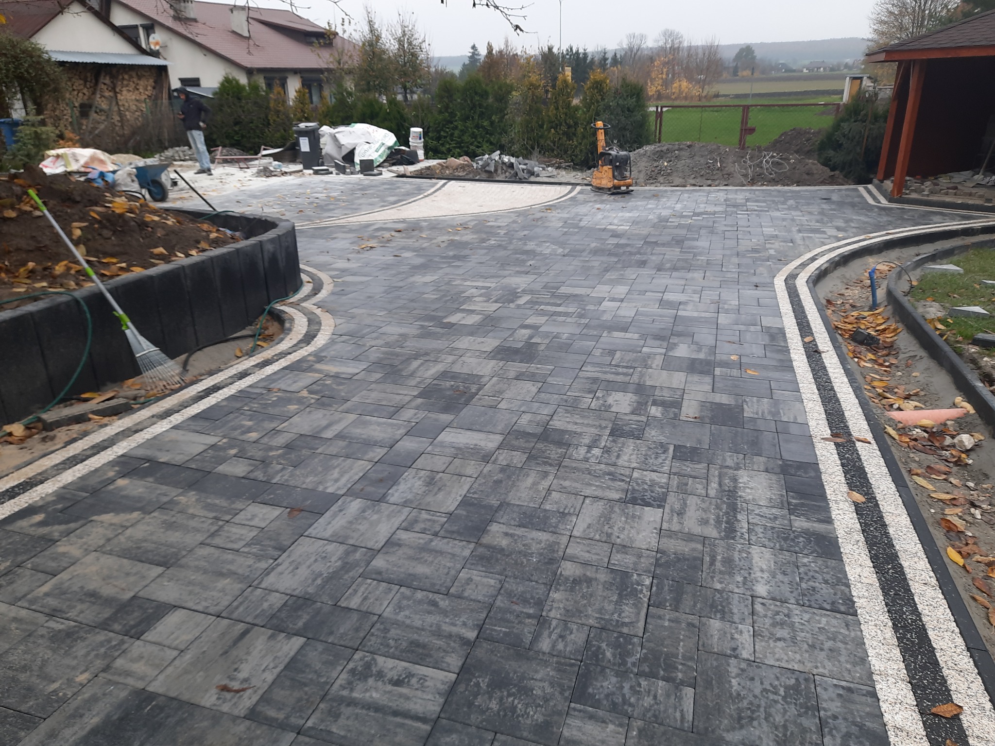 Ułożona szara kostka brukowa z jasnymi pasami oddzielającymi, widoczny fragment ogrodu, w tle dom i pracownik budowlany, na placu budowy stoi zagęszczarka.