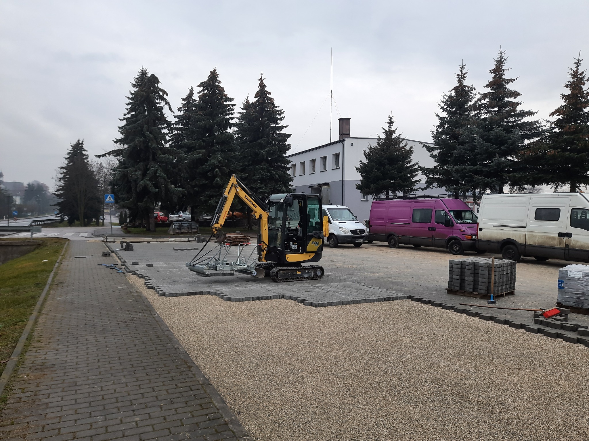 Żółta mini-koparka z przyssawkami do kostki brukowej w trakcie pracy na podsypce żwirowej, ułożone fragmenty nawierzchni z szarej kostki, w tle zaparkowane busy i budynek.