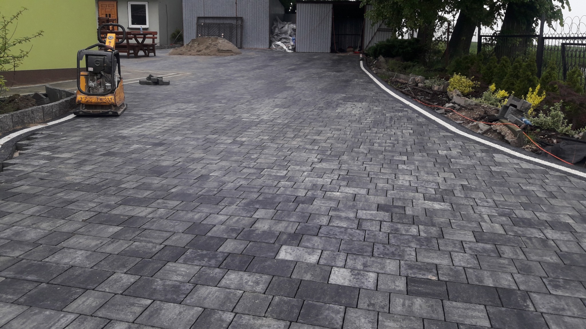 Ułożona szara kostka brukowa na podjeździe, widoczna zagęszczarka wibracyjna oraz obrzeża z kostki granitowej oddzielające nawierzchnię od trawnika.