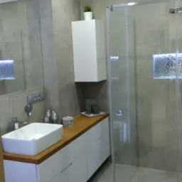 Nowoczesna łazienka z szarymi płytkami, białą szafką pod umywalką z drewnianym blatem, kabiną prysznicową ze szklanymi ścianami i dekoracyjną mozaiką w ścianie.