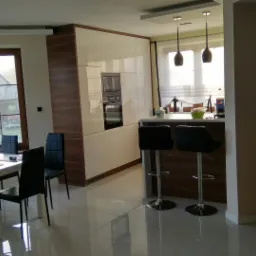 Jasna kuchnia z białymi szafkami i drewnianymi akcentami, wysoki blat z hokerami, stół z krzesłami przy oknie, nowoczesne lampy wiszące.