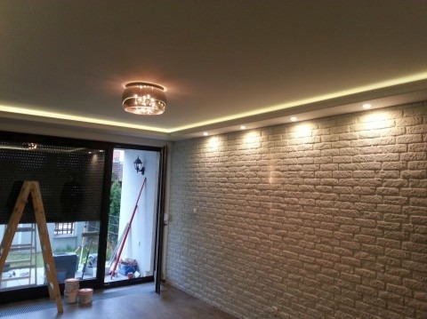 Pokój po odświeżeniu z dekoracyjną ścianą z cegły, oświetleniem LED podsufitowym i nowoczesną lampą wiszącą, widoczna drabina malarska i puszki z farbą.