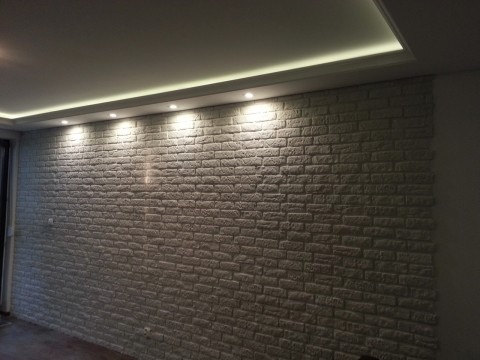 Ściana wykończona dekoracyjną białą cegłą, podświetlona punktowymi lampami sufitowymi i listwą LED, tworząca nowoczesny efekt wnętrza.