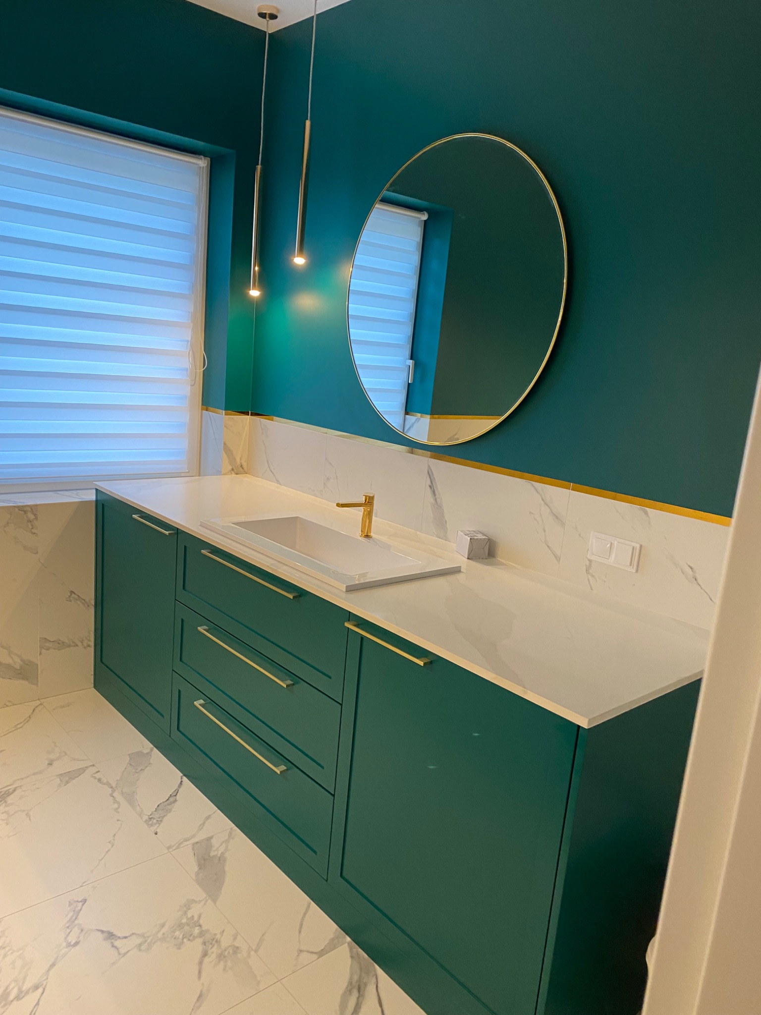 Nowoczesna łazienka z zielonymi szafkami pod umywalką, białym blatem i marmurowymi płytkami na podłodze i ścianie, okrągłe lustro z złotą ramą i wiszące lampy.