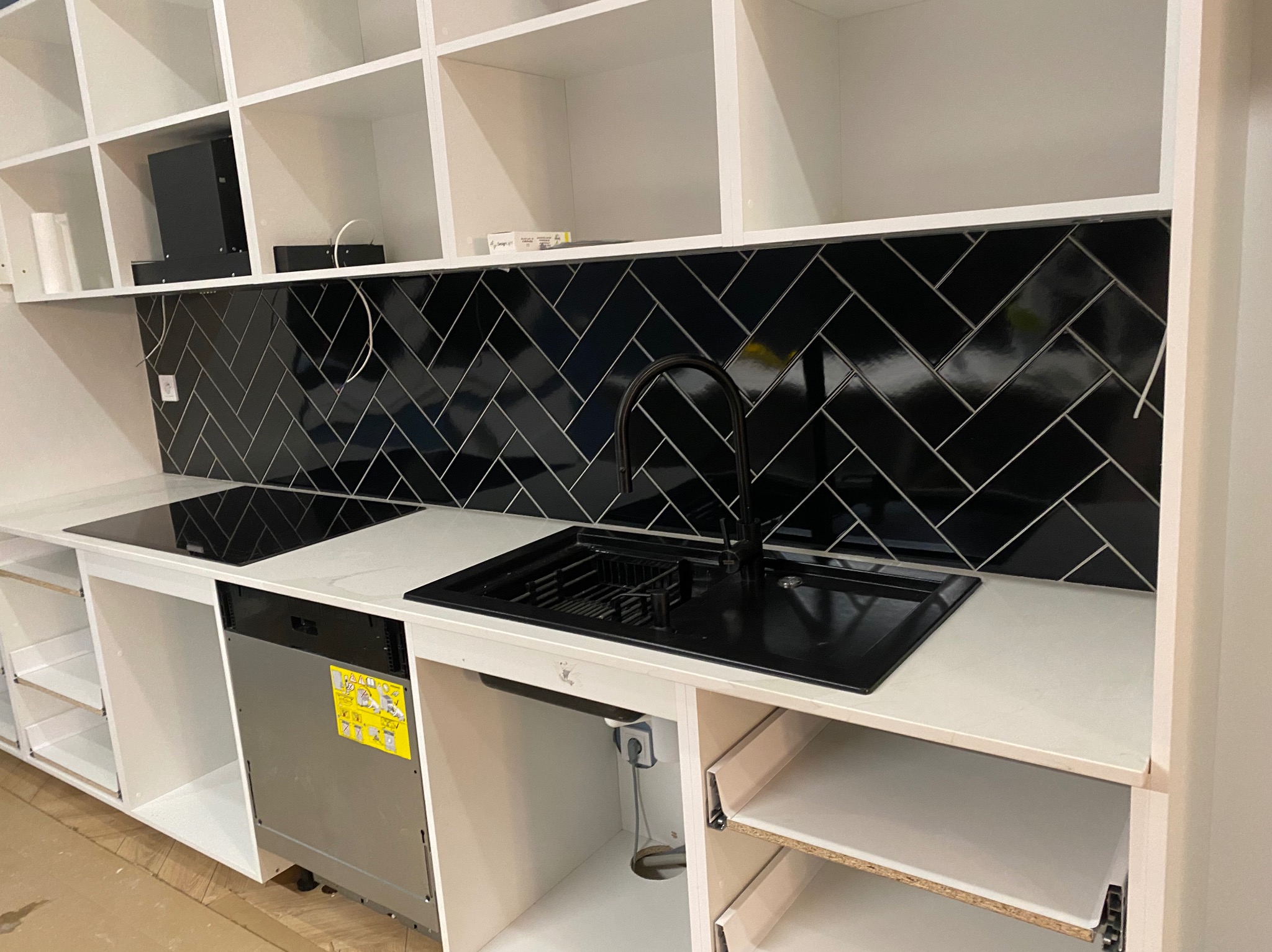Nowoczesna kuchnia z czarną armaturą, czarnymi płytkami ułożonymi w jodełkę na ścianie i białymi szafkami, z widoczną zmywarką z naklejkami informacyjnymi i częściowo zamontowanymi szufladami.