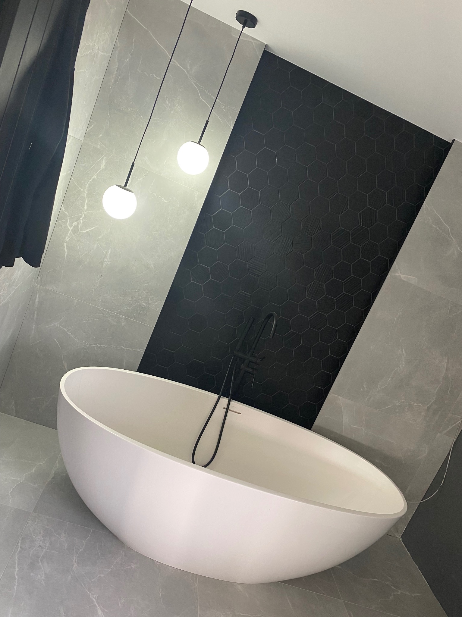Nowoczesna łazienka z wolnostojącą białą wanną, czarną armaturą i szarymi płytkami imitującymi marmur, z akcentem czarnej ściany z heksagonalnymi płytkami i wiszącymi lampami kulami.