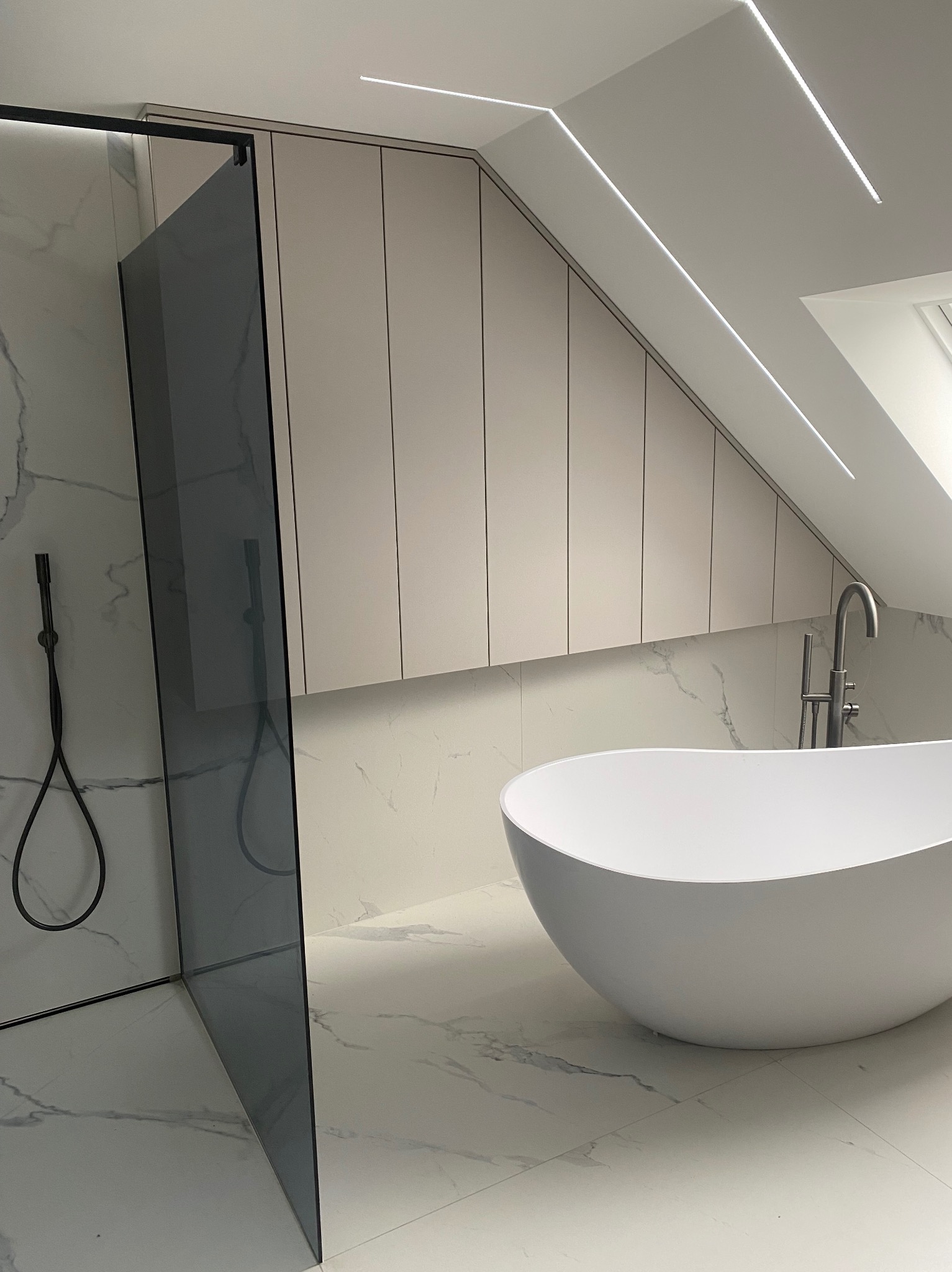 Nowoczesna łazienka na poddaszu z wolnostojącą wanną, szklaną kabiną prysznicową z czarnym wykończeniem, zabudową meblową pod skosem i marmurowymi płytkami.