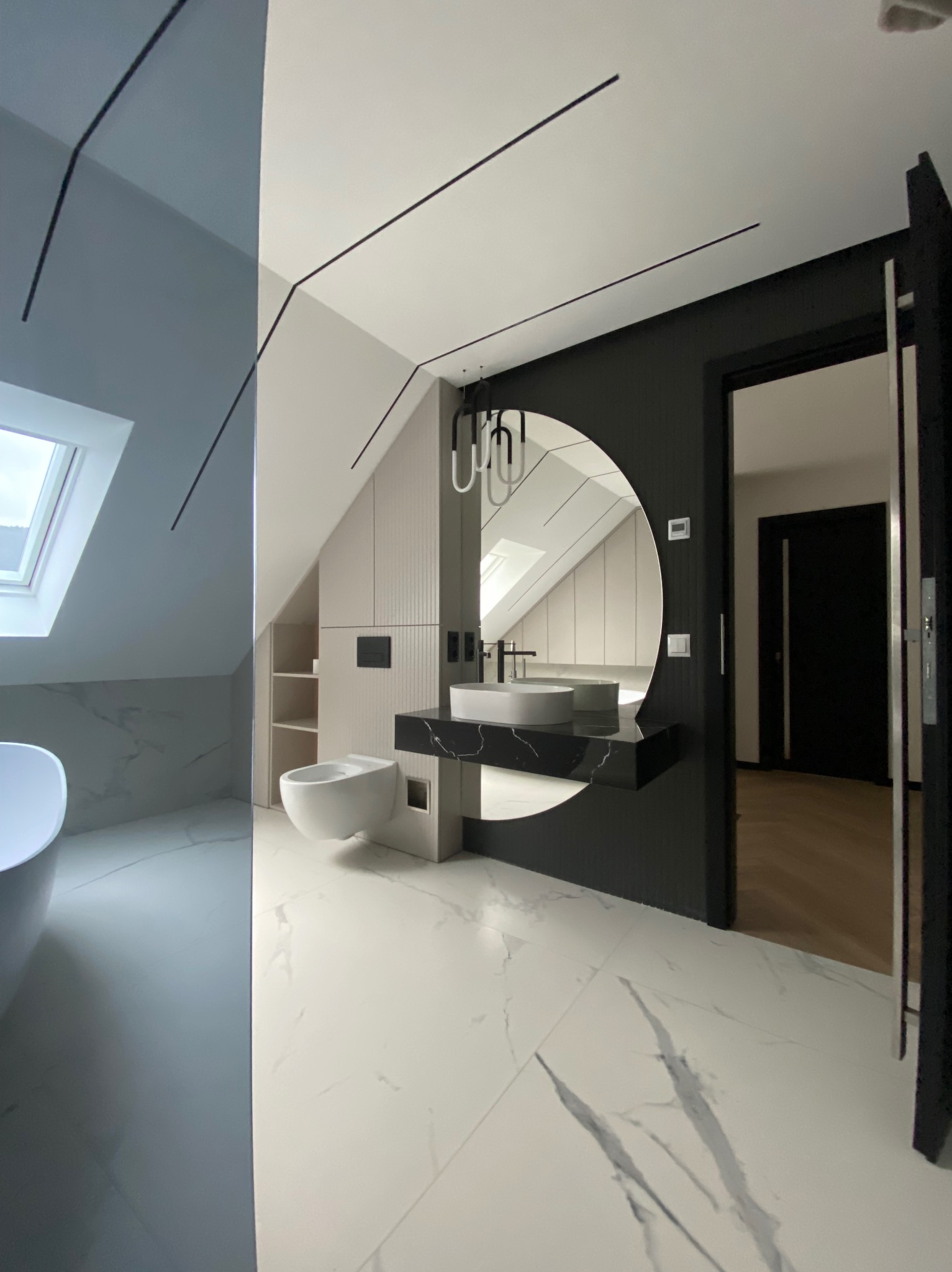Nowoczesna łazienka na poddaszu z białą armaturą, umywalką na czarnym marmurowym blacie, okrągłym lustrem i płytkami imitującymi marmur na podłodze i części ścian.