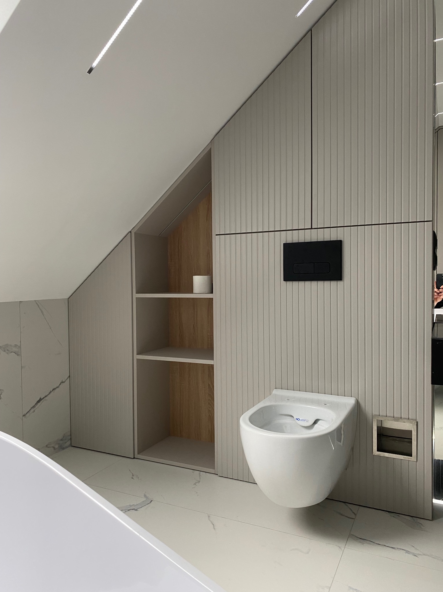 Nowoczesna łazienka na poddaszu ze ścianą z pionowymi panelami, podwieszaną miską WC z czarnym przyciskiem spłukującym i otwartą szafką z półkami.