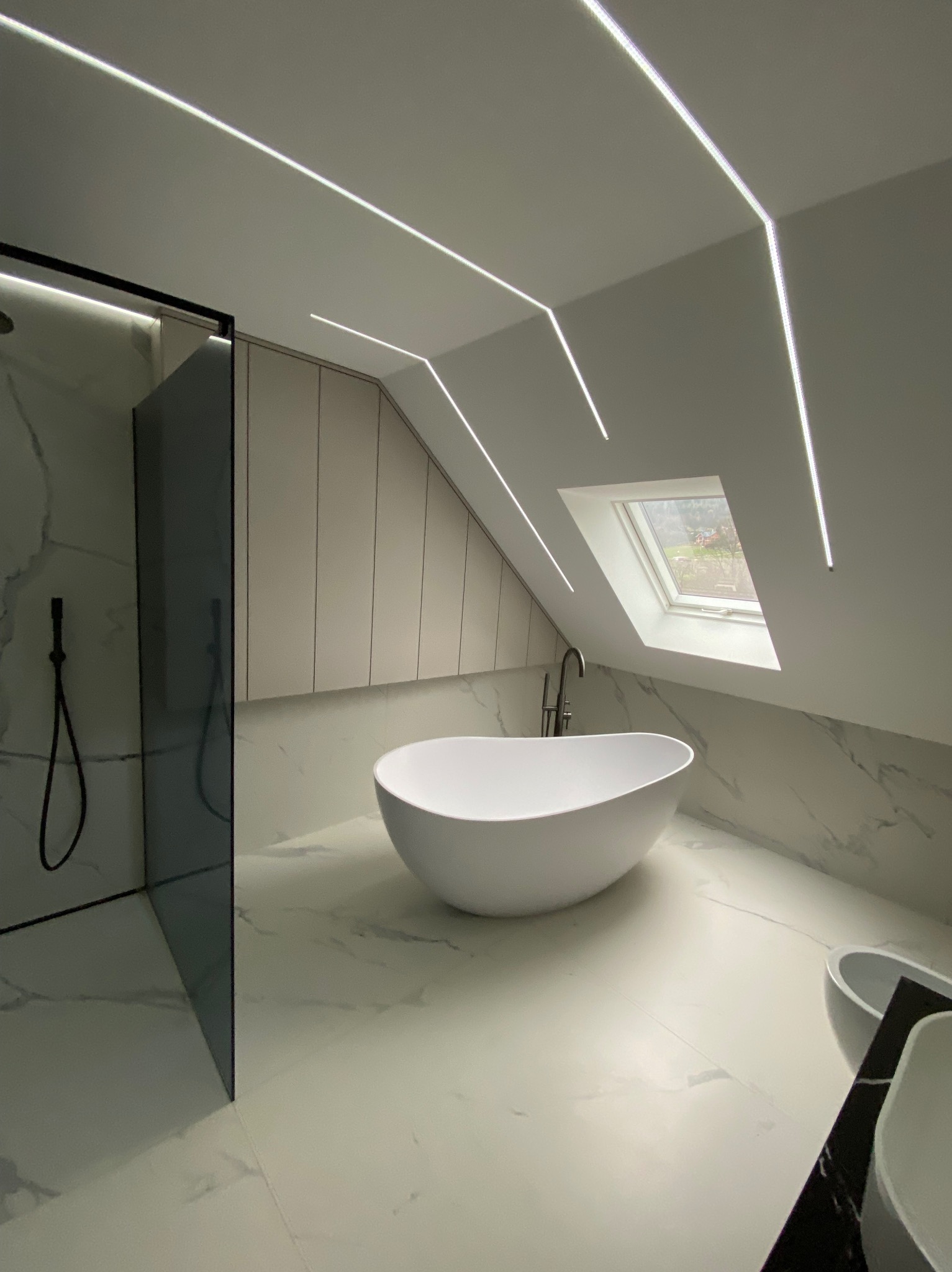 Nowoczesna łazienka na poddaszu z białą wanną wolnostojącą, kabiną prysznicową ze szkła w czarnej ramie, marmurowymi płytkami, oknem dachowym i liniowym oświetleniem LED.