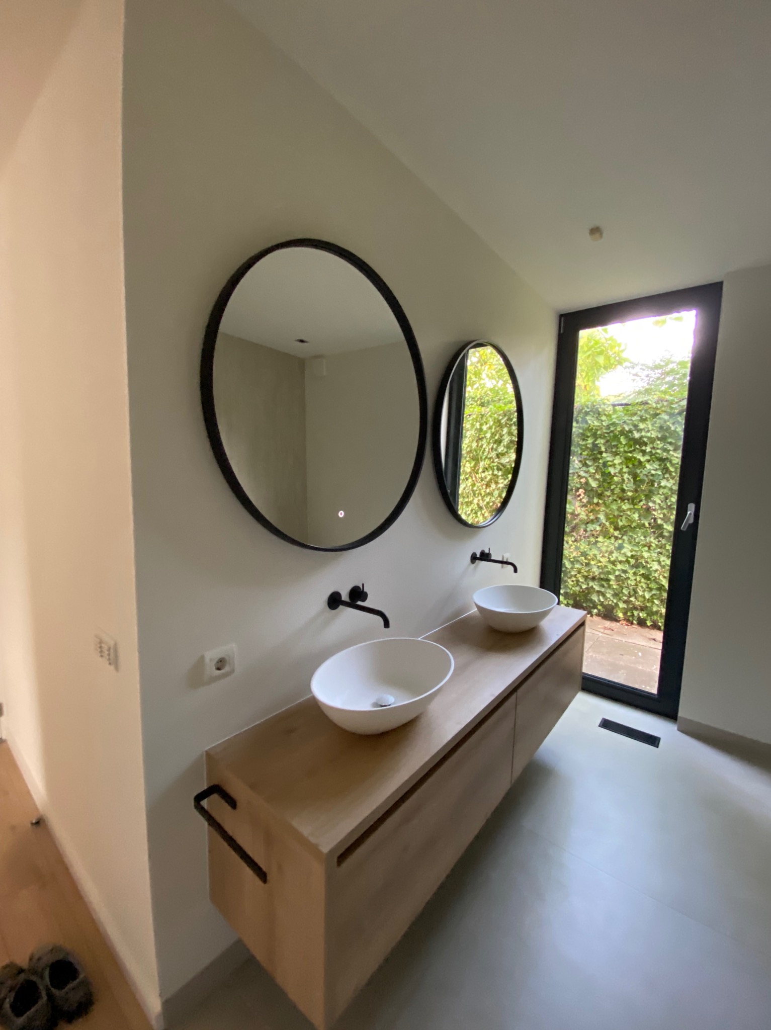 Nowoczesna łazienka z dwoma okrągłymi lustrami w czarnych ramach, wiszącą szafką z drewnianym blatem, białymi umywalkami nablatowymi i czarną armaturą na tle okna z widokiem na zielony żywopłot.