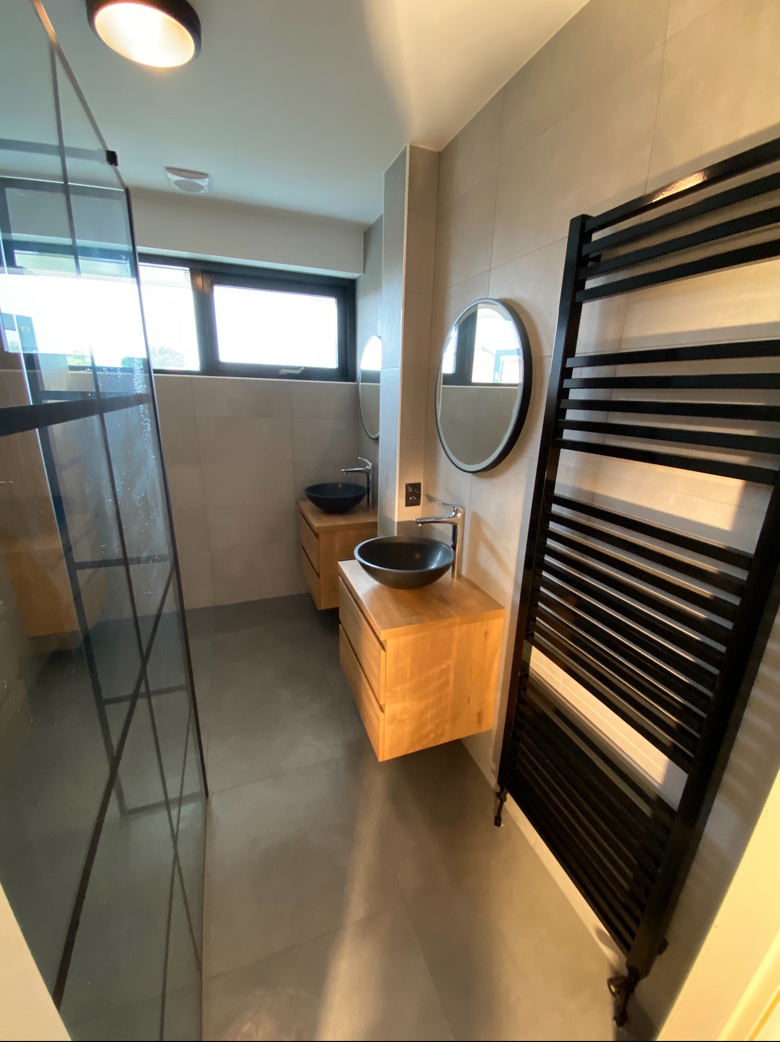 Nowoczesna łazienka z drewnianymi szafkami pod umywalki z czarnymi misami, okrągłe lustra, czarny grzejnik drabinkowy i kabina prysznicowa ze szklaną ścianką z czarnymi profilami.