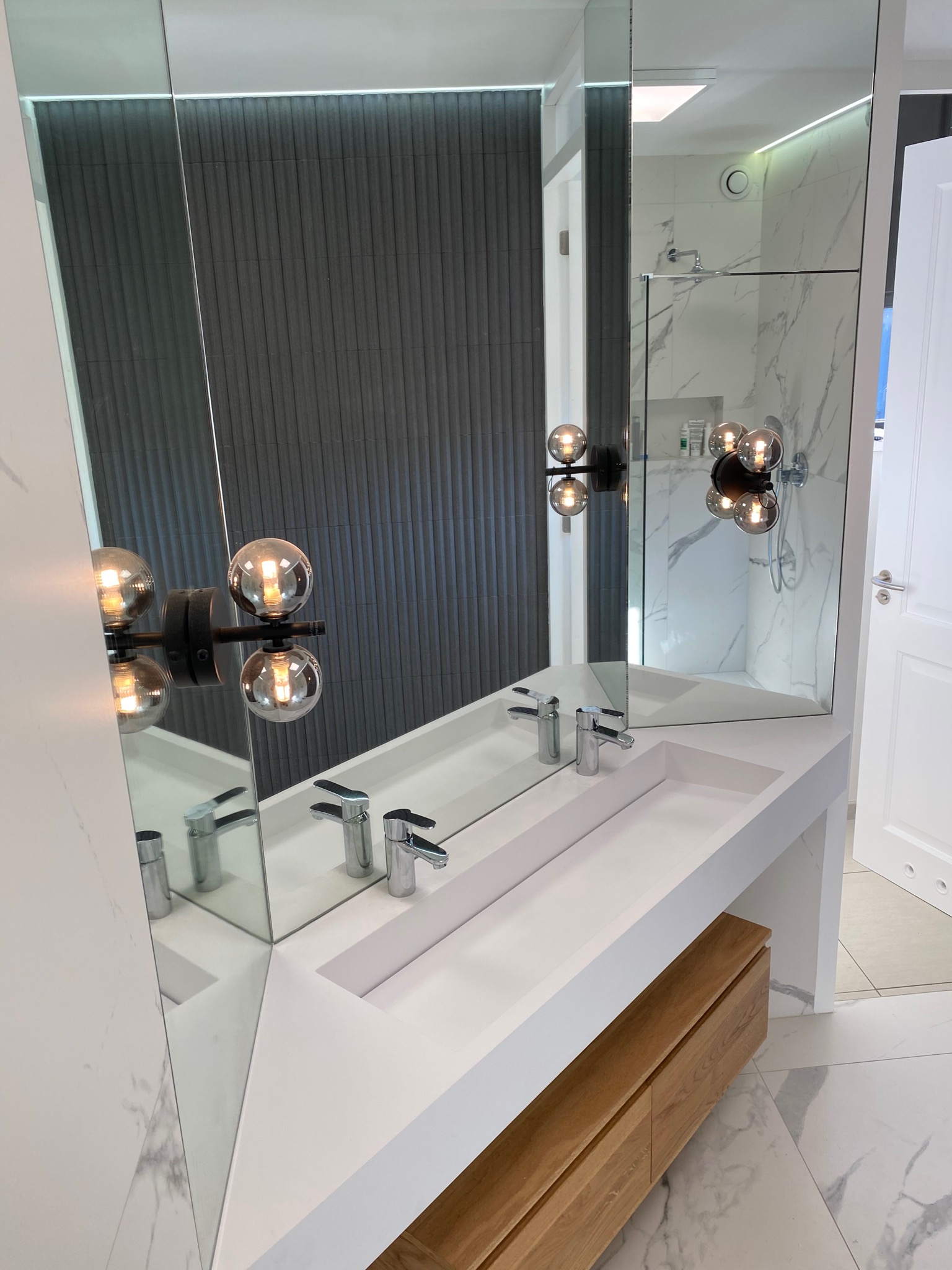 Nowoczesna łazienka z podwójną umywalką wpuszczoną w biały blat, drewniana szafka pod blatem, duże lustro z dekoracyjnymi lampami kulami, marmurowe płytki na ścianach w kabinie prysznicowej.