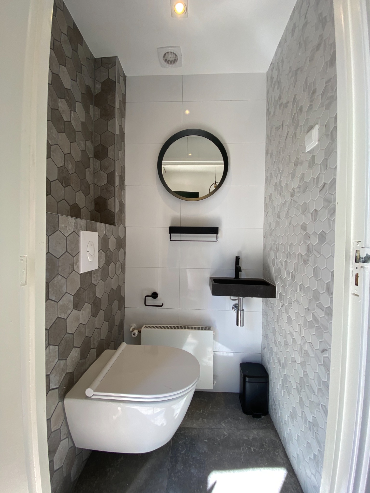 Nowoczesna toaleta z heksagonalnymi płytkami w odcieniach szarości, wisząca muszla klozetowa, czarna umywalka nablatowa i okrągłe lustro z czarną ramą.