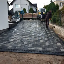 Ułożona kostka brukowa w odcieniach szarości na posesji, widoczny pracownik z wiaderkiem, krawężnik z odpływem liniowym i samochód ciężarowy w tle.