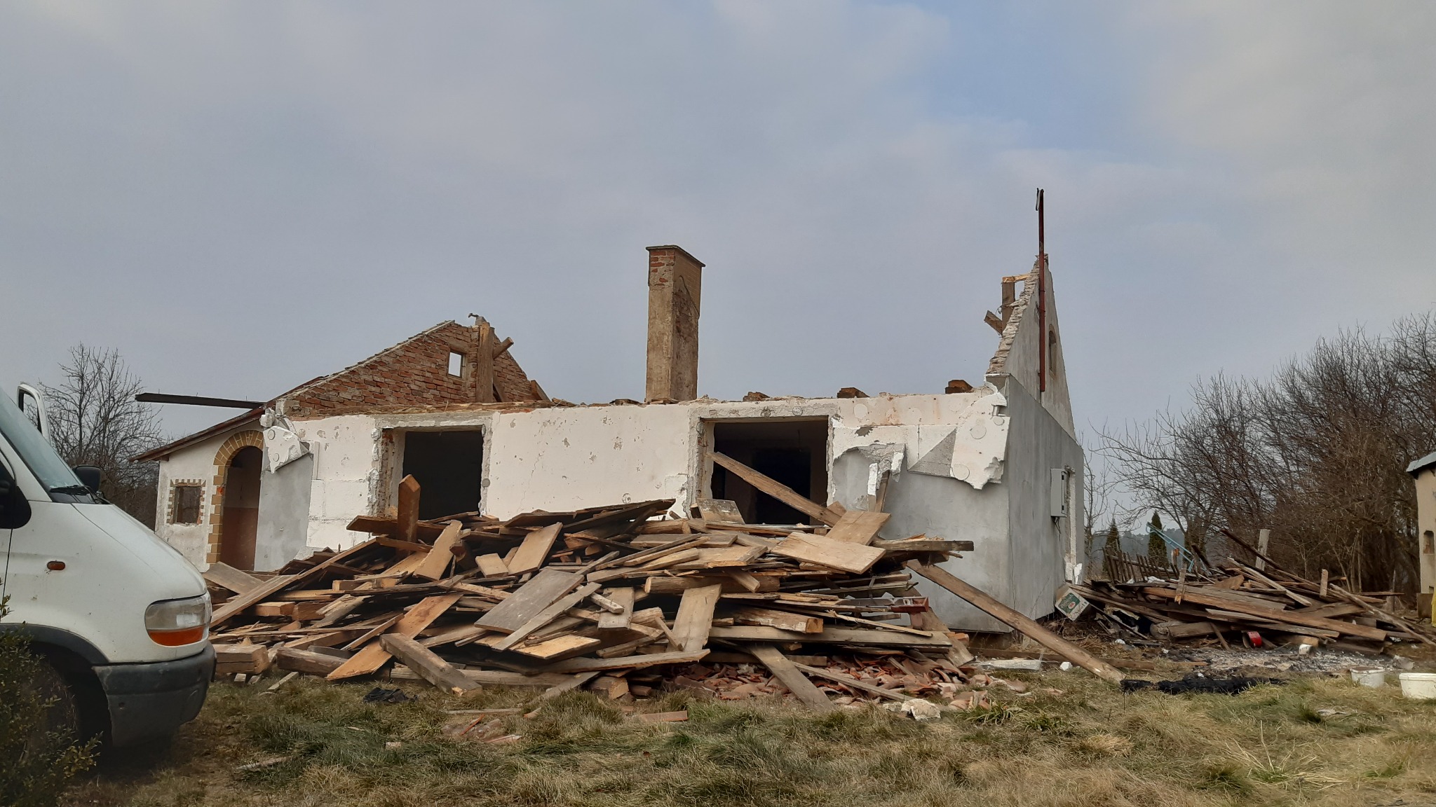 Częściowo rozebrany budynek z fragmentami ścian i komina w Olsztynie, z kupą gruzu przed nim i białym samochodem dostawczym z otwartymi drzwiami po lewej stronie kadru.
