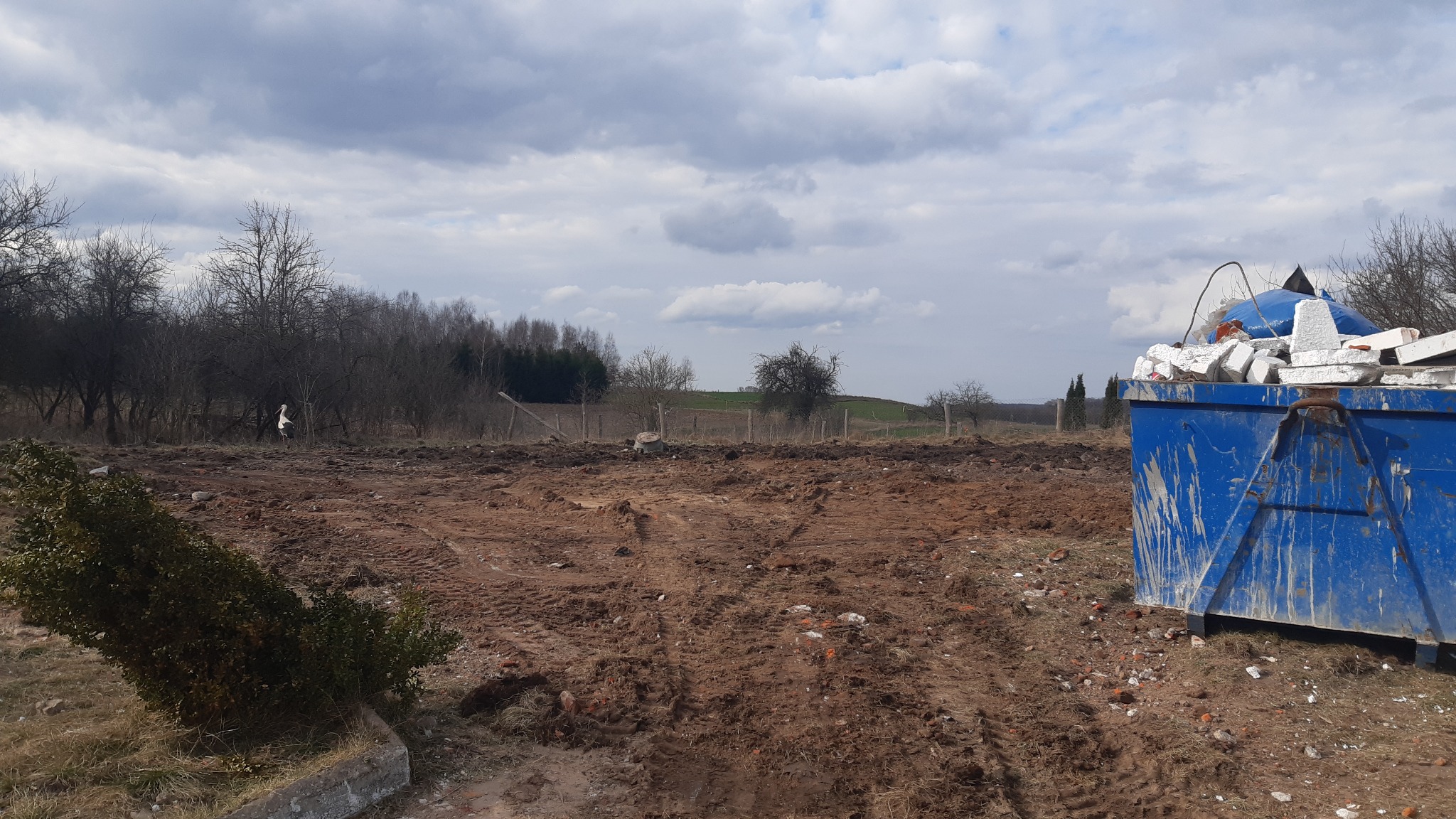 Nierówny teren po pracach ziemnych z widocznym, wypełnionym gruzem, niebieskim kontenerem, w tle drzewa i pochmurne niebo.