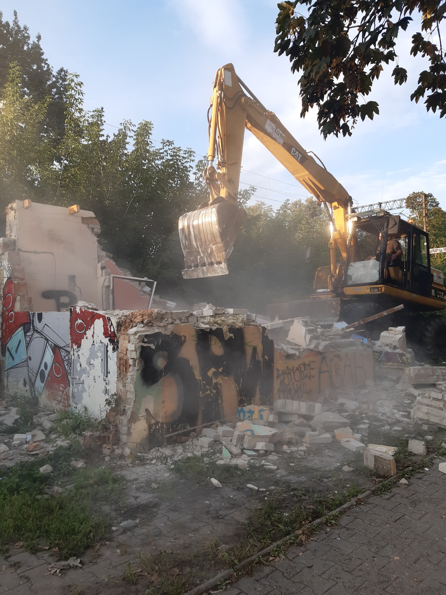 Żółta koparka Caterpillar w trakcie rozbiórki częściowo zburzonego budynku pokrytego graffiti, unoszący się pył i gruz, widoczne drzewa w tle.
