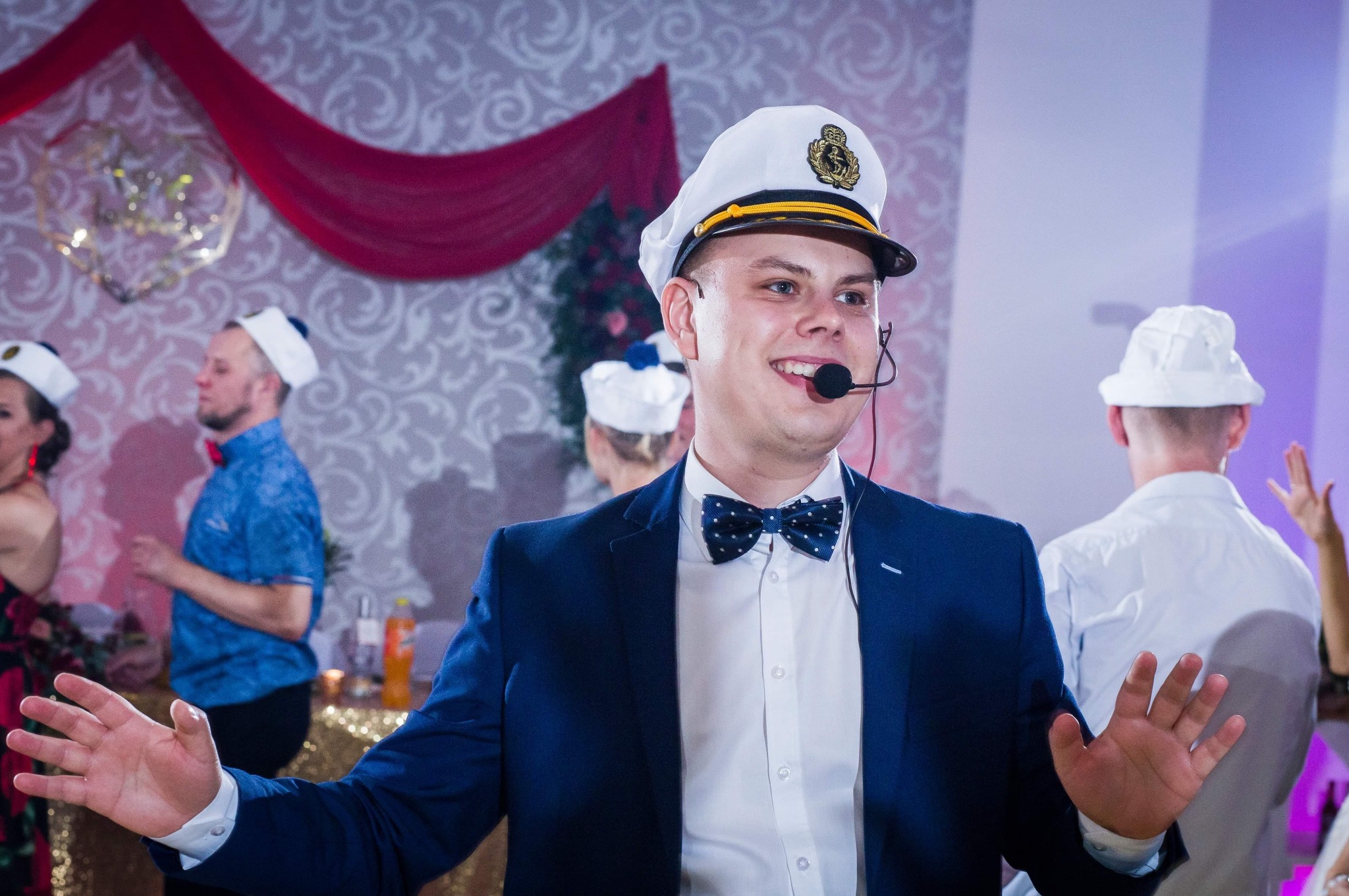 Uśmiechnięty konferansjer w marynarskiej czapce, muszce i mikrofonem nagłownym, gestykulujący podczas imprezy z motywem morskim, w tle zespół w marynarskich czapkach i dekoracje.