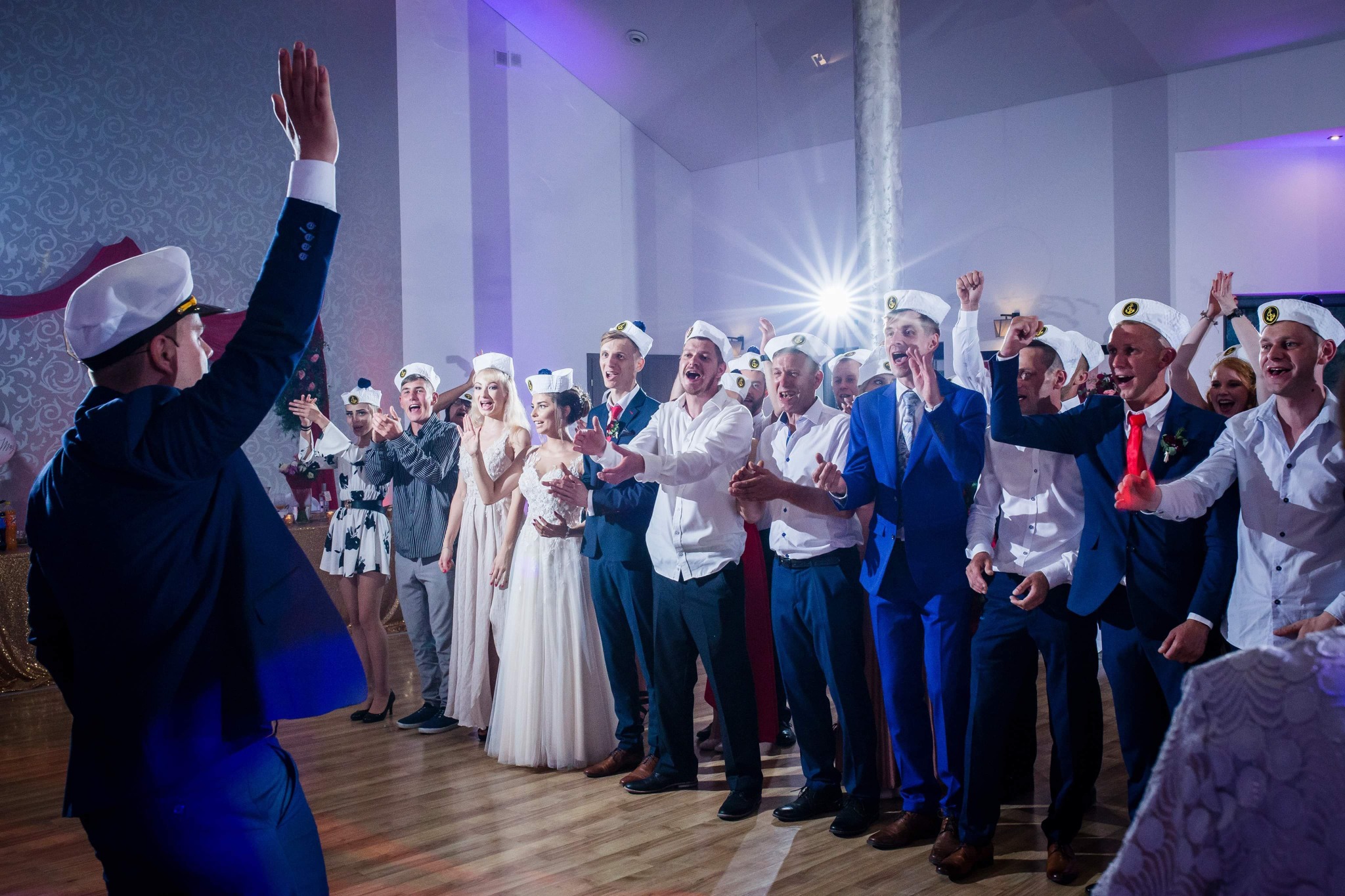 Grupa gości weselnych w marynarskich czapkach, ustawiona w rzędzie i klaszcząca podczas uroczystości, oświetlenie sceniczne w tle.