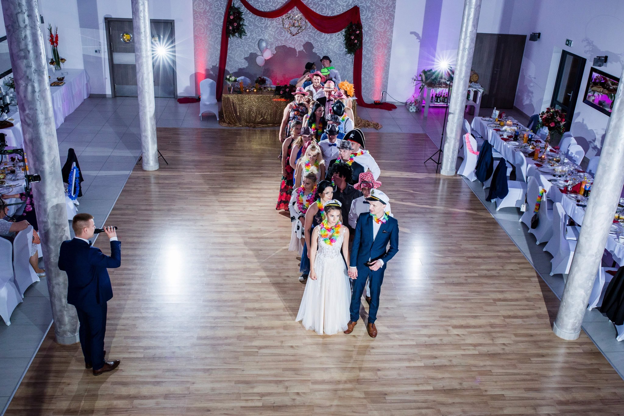 Para młoda prowadzi korowód gości weselnych w przebraniach przez salę bankietową z kolumnami i dekoracjami.