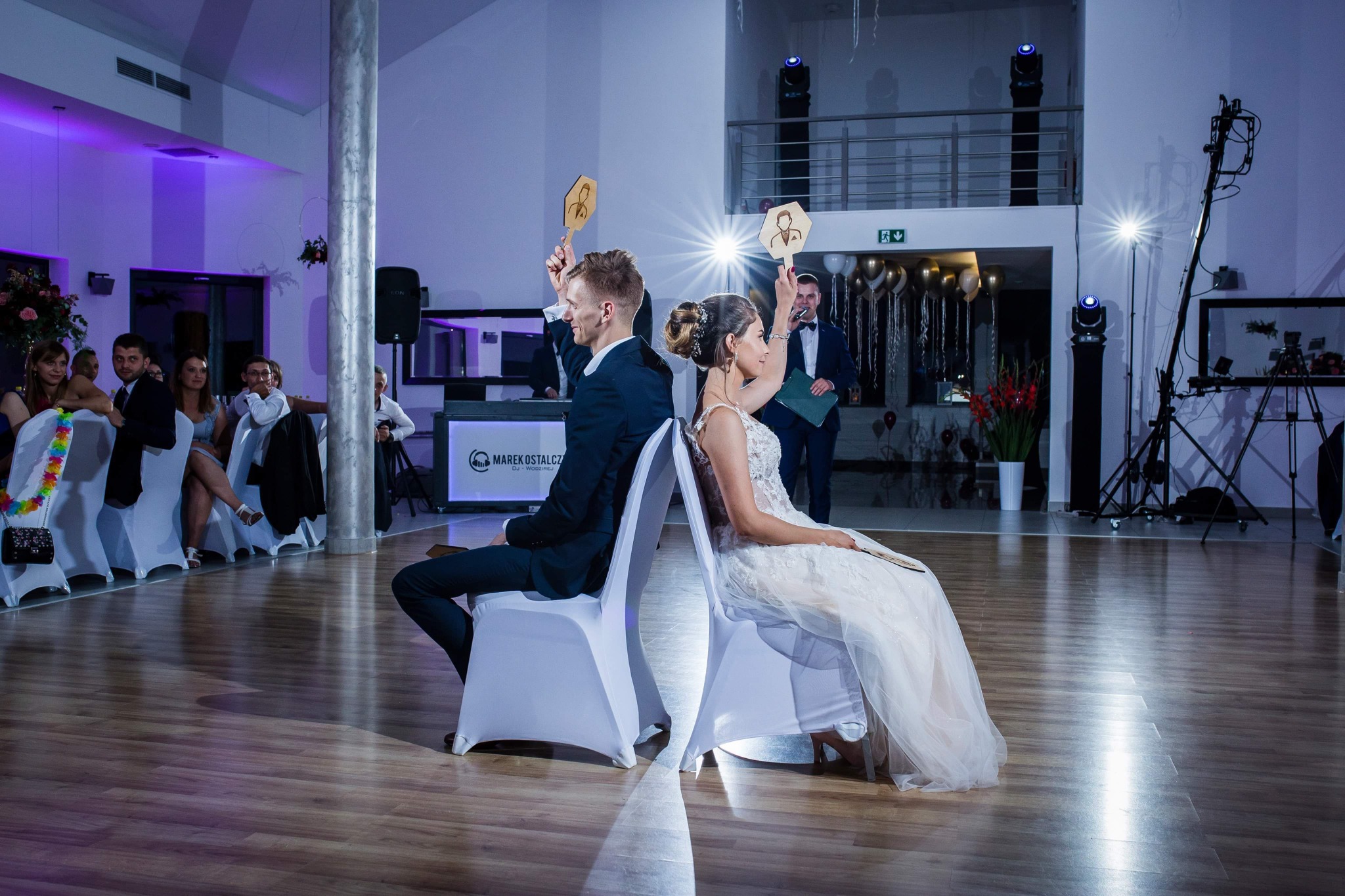 Para młoda siedząca tyłem do siebie na krzesłach, trzymająca w górze tabliczki z wizerunkami, podczas oczepin na sali weselnej z dekoracjami i oświetleniem.