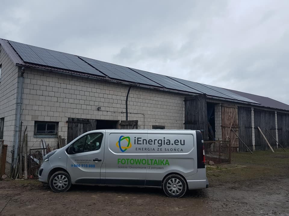 Szary bus firmowy z logo i napisem 'FOTOWOLTAIKA' zaparkowany przed budynkiem gospodarczym z panelami słonecznymi na dachu, pochmurne niebo w tle.