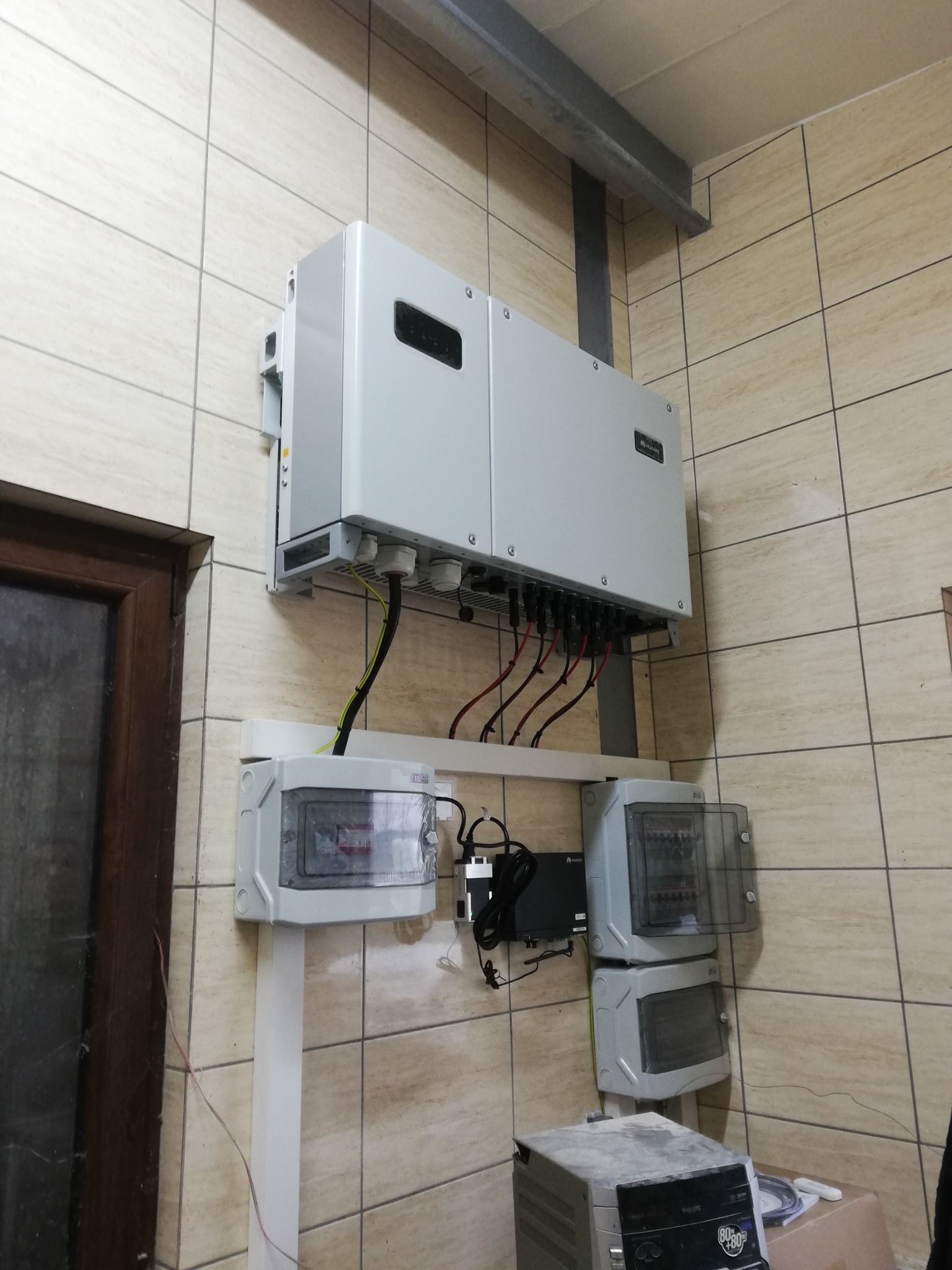 Zamontowany inwerter fotowoltaiczny wraz z rozdzielnicą i okablowaniem na kafelkowanej ścianie w Olsztynie. Widoczne złącza i przewody elektryczne.