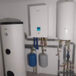 Biała kotłownia z trzema zbiornikami (jeden pionowy, dwa poziome), szarymi rurami i niebieskimi elementami, zamontowana na tle białych płytek.