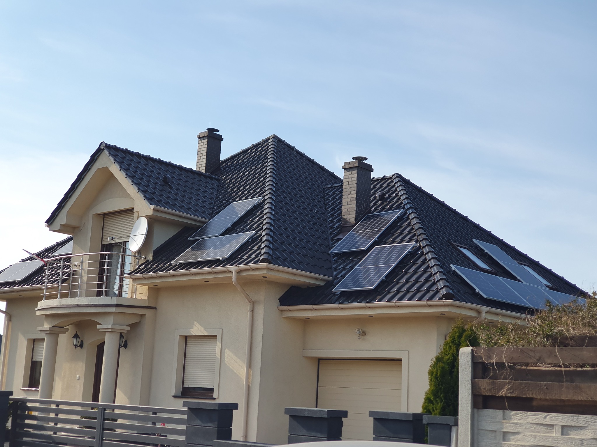 Dwukondygnacyjny dom jednorodzinny z ciemną dachówką i panelami słonecznymi na dachu, widoczne dwa kominy, balkon z metalową balustradą i częściowo zasłonięte okna roletami.