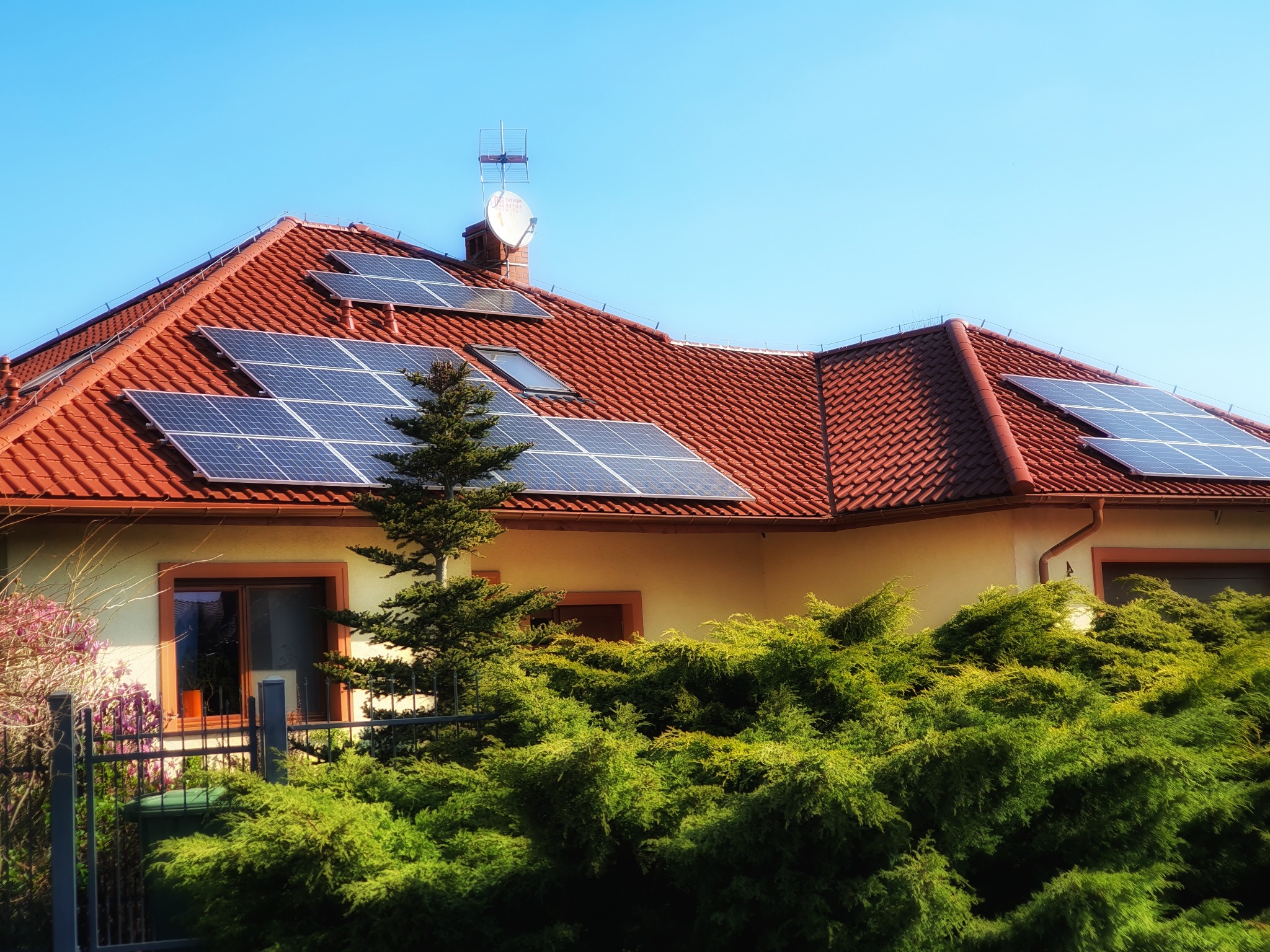 Dom jednorodzinny z czerwoną dachówką i panelami słonecznymi na dachu, antena satelitarna na kominie, bujna roślinność w ogrodzie.