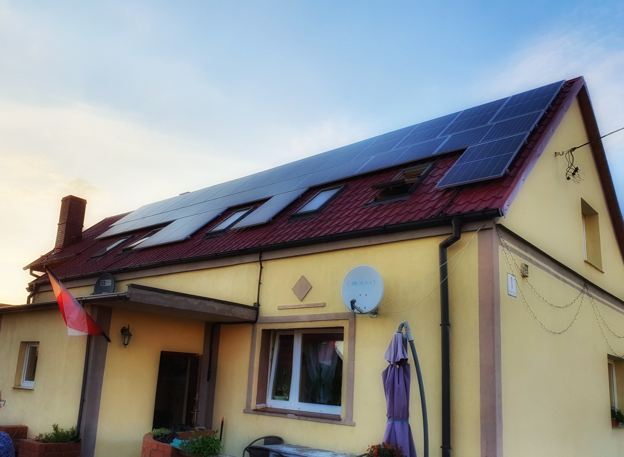 Żółty dom jednorodzinny w Legnicy z panelami słonecznymi na czerwonym dachu i oknami dachowymi, widziany z dołu pod kątem, z polską flagą przy wejściu.