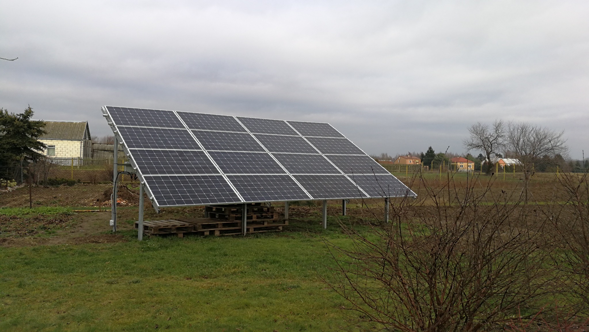 Instalacja fotowoltaiczna na metalowej konstrukcji, panele ustawione pod kątem, w tle zabudowania wiejskie i pochmurne niebo.