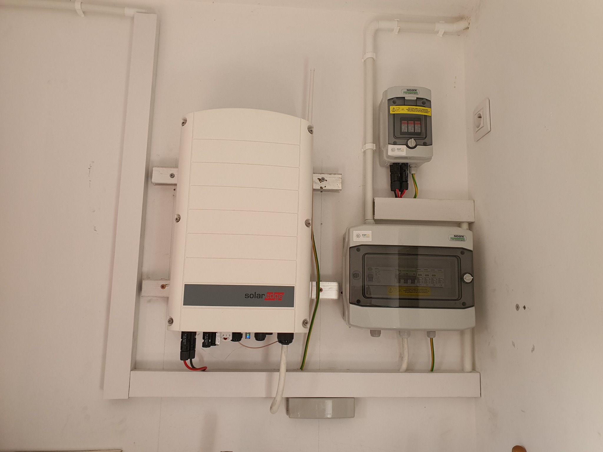 Biały inwerter SolarEdge z szarą listwą, zamontowany na ścianie obok skrzynki bezpiecznikowej Noark wewnątrz budynku.