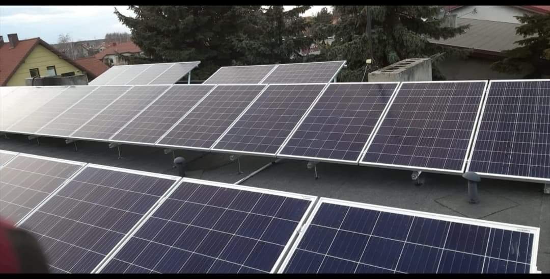 Rząd paneli słonecznych na płaskim dachu z widocznymi drzewami i domami w tle, pochmurne niebo.