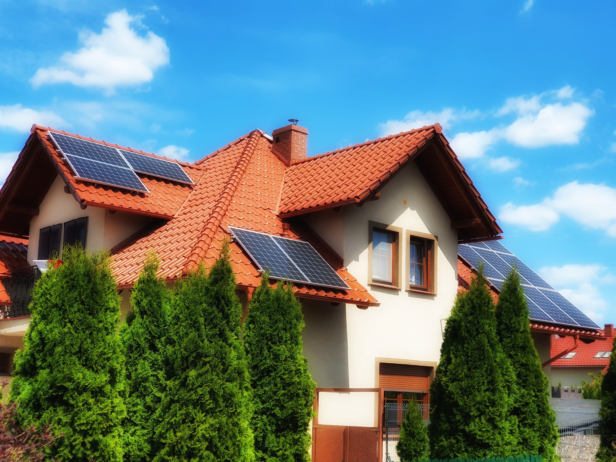 Dom jednorodzinny z panelami słonecznymi na czerwonym dachu, widok z poziomu ogrodu z zielonymi tujami, błękitne niebo z chmurami.