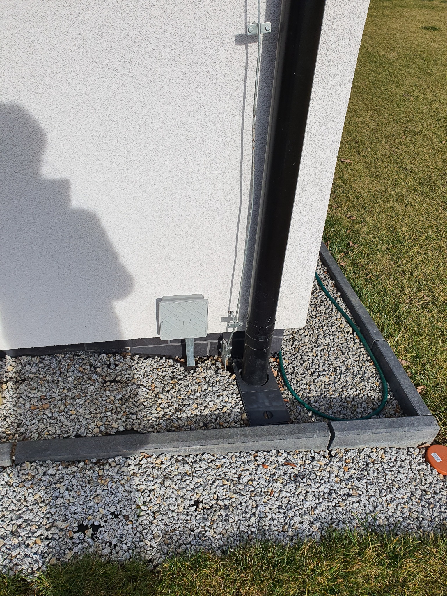 Czarny pionowy przewód instalacji przy białej ścianie budynku, widoczne elementy mocujące, skrzynka instalacyjna, kamyki i trawa na gruncie.