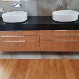 Łazienka z dwoma owalnymi umywalkami nablatowymi na czarnym, błyszczącym blacie z szafką z jasnego drewna z czterema szufladami i srebrnymi uchwytami.