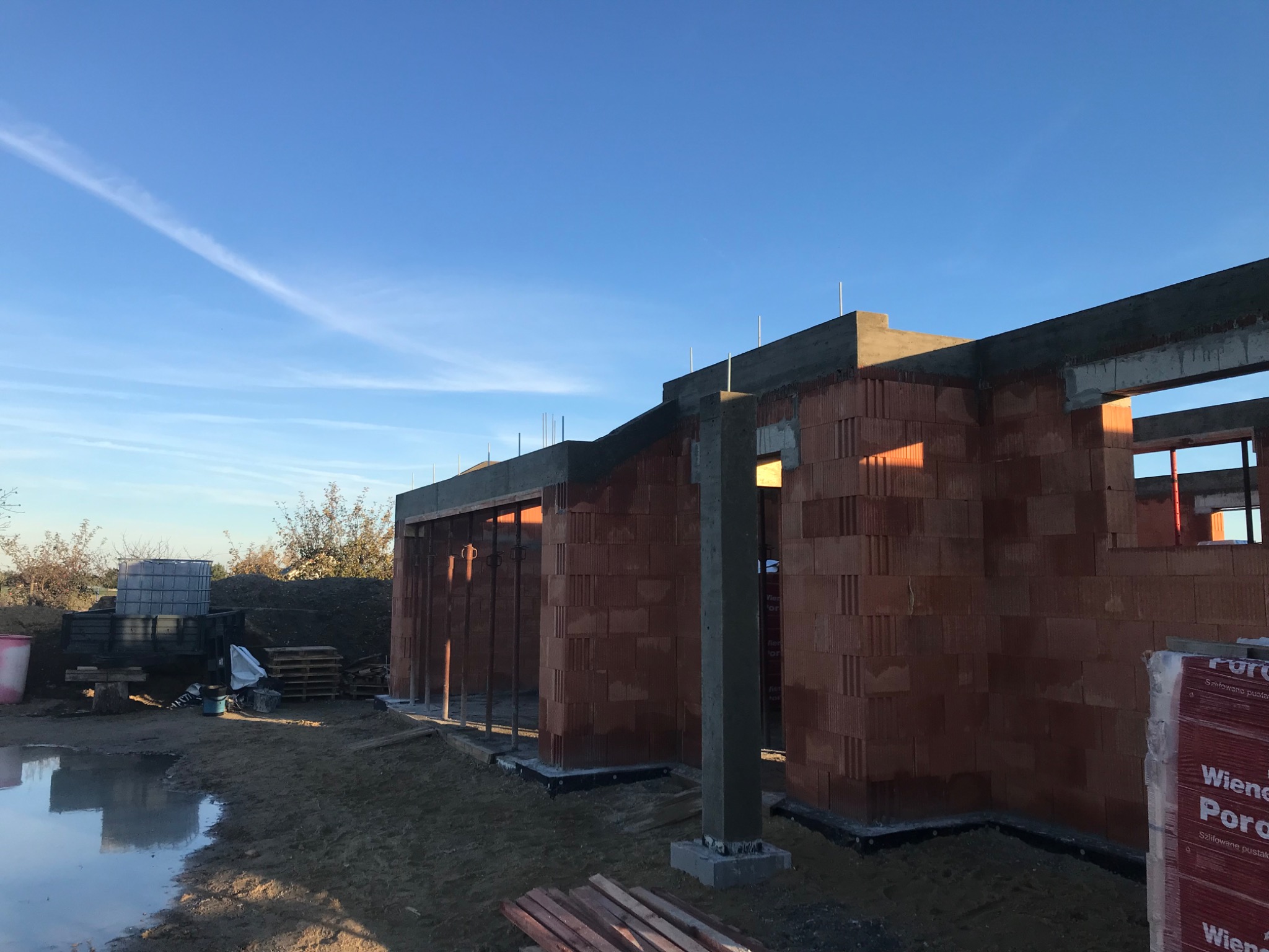 Wznoszenie ścian budynku z czerwonej cegły, widoczne zbrojenia i betonowe nadproża, teren budowy z kałużą i materiałami budowlanymi, błękitne niebo w tle.