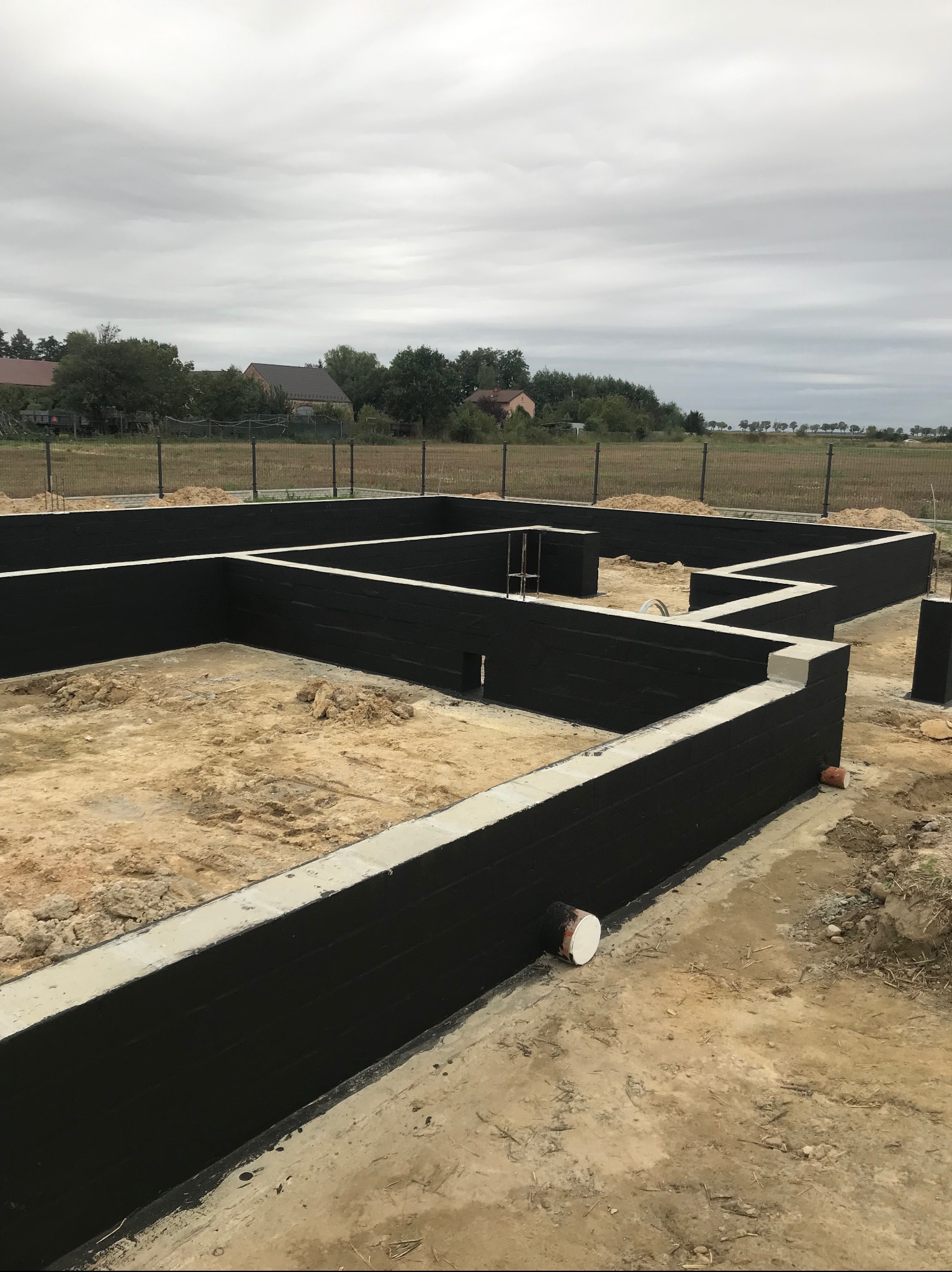 Zabezpieczone fundamenty domu jednorodzinnego, ściany pomalowane na czarno, widoczne rury kanalizacyjne, ziemia wewnątrz, pochmurne niebo w tle.