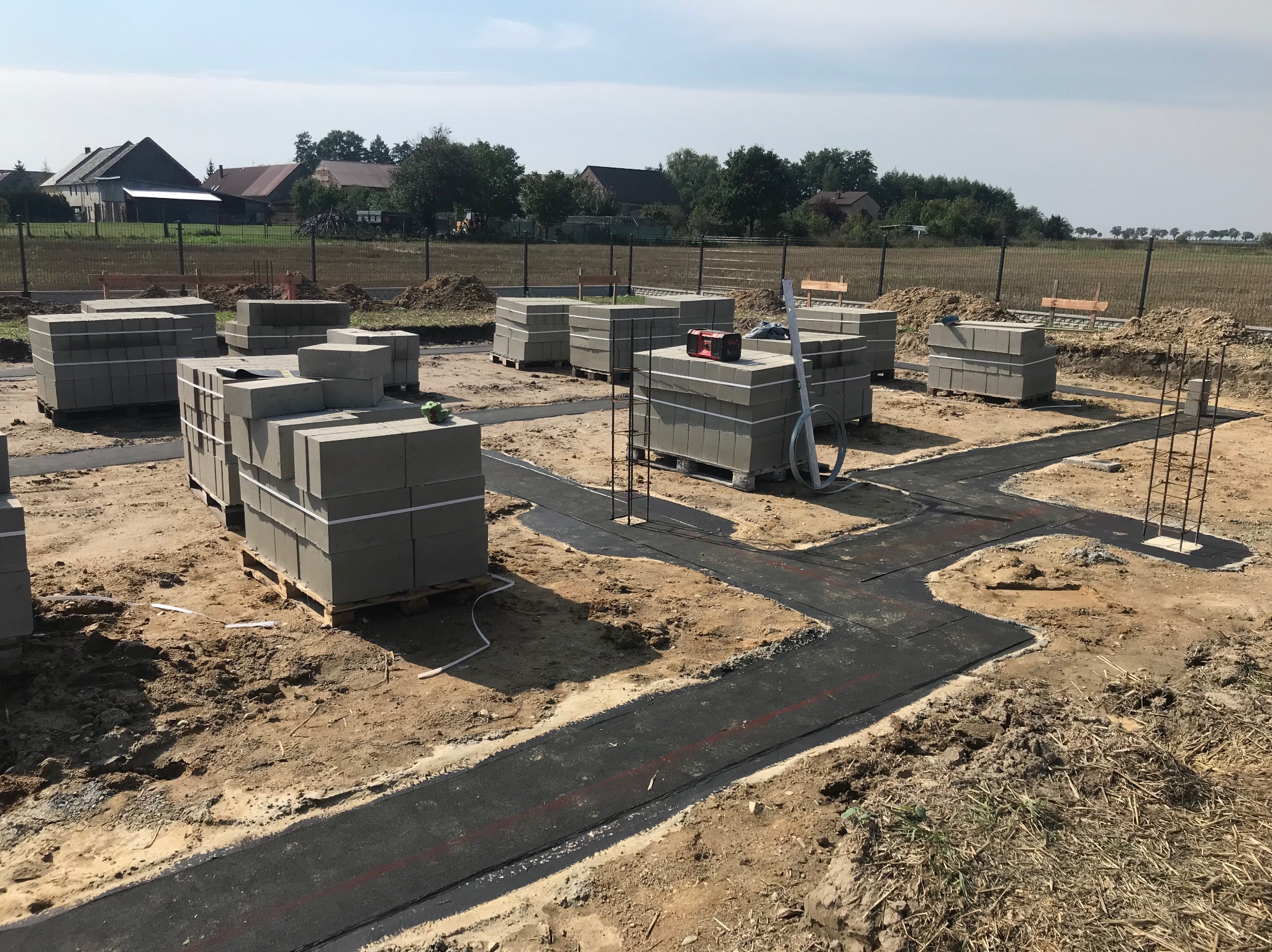 Przygotowanie fundamentów: ułożona izolacja przeciwwilgociowa w wykopie, palety z bloczkami betonowymi, zbrojenie. Widok na teren budowy i zabudowania w tle.