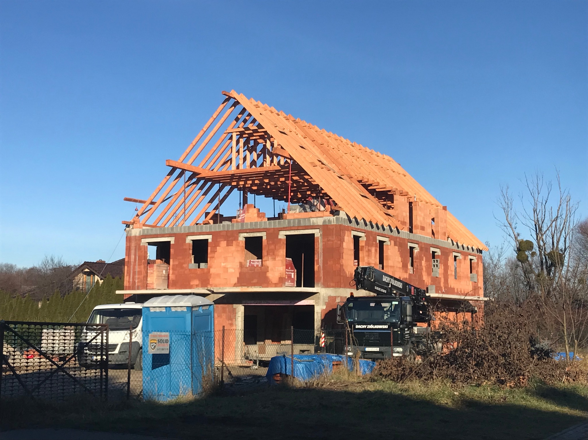 Budowa domu z czerwonej cegły w trakcie krycia dachu dachówką, widoczny dźwig i przenośna toaleta. Konstrukcja dachu z drewnianych belek na tle błękitnego nieba.