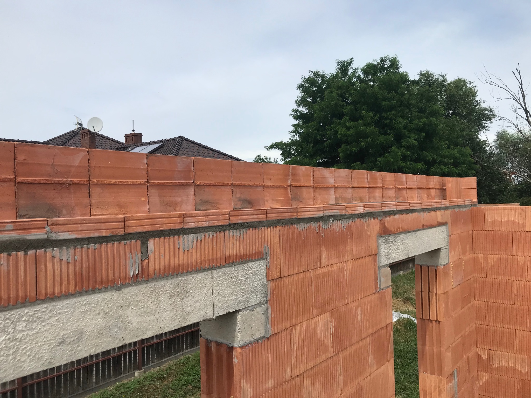 Wznoszone ściany z czerwonej cegły ceramicznej, widoczne betonowe nadproża okienne. W tle zieleń drzew i fragment dachu budynku. Etap budowy domu.