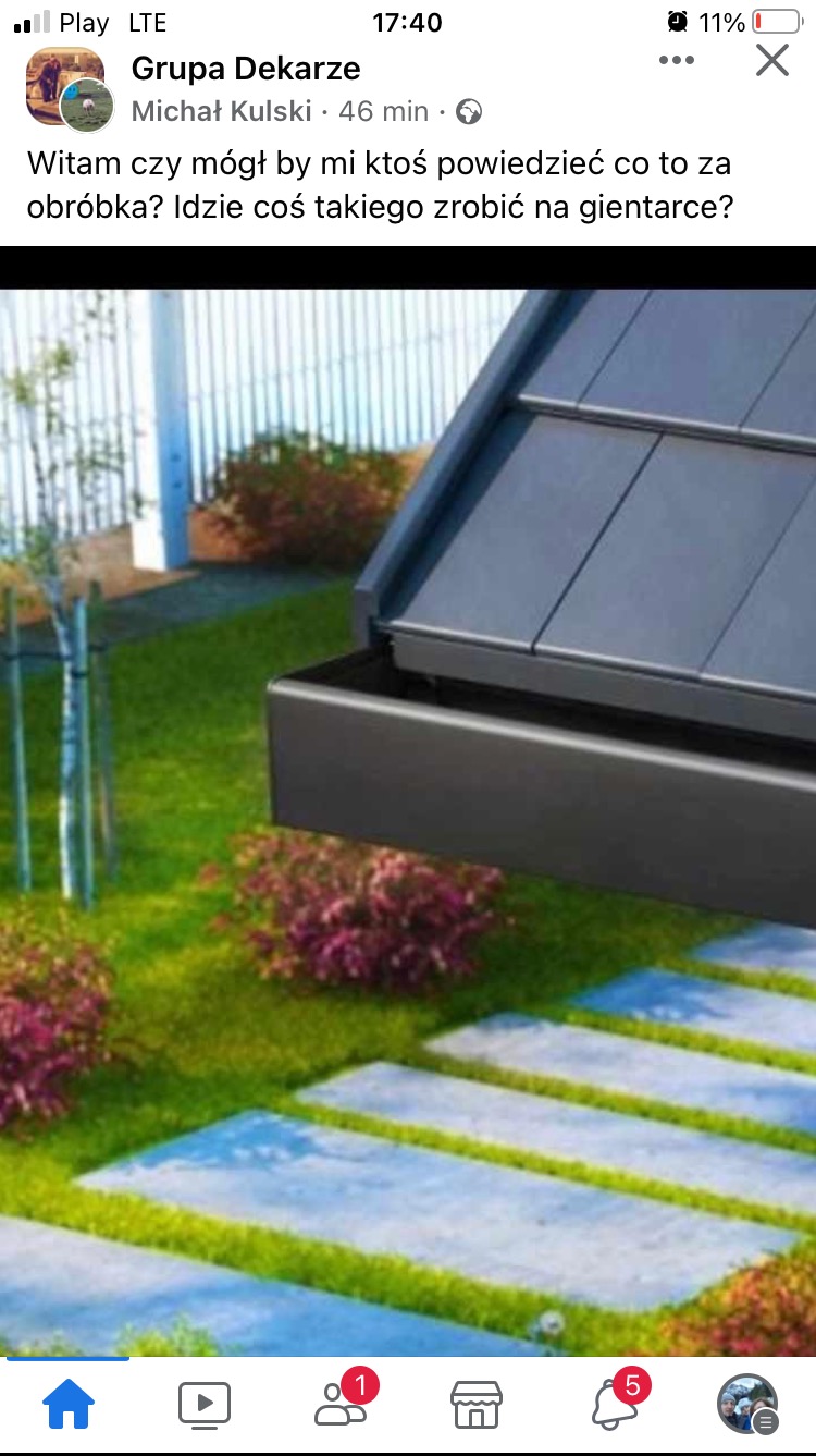 Szara rynna dachowa z fragmentem dachu na tle zielonego trawnika i ścieżki z płyt. Ujęcie z dołu, prezentujące detale obróbki blacharskiej.