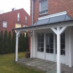 Zadaszenie nad tarasem z białymi, drewnianymi słupami i szarą, spadzistą konstrukcją dachu, przylegające do ceglanego domu z białymi drzwiami balkonowymi, widok na trawnik i rząd tuj.