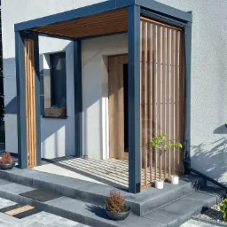 Nowoczesna pergola przy wejściu do domu z szarymi elementami konstrukcyjnymi i drewnianymi lamelami, z małymi roślinami w doniczkach po bokach wejścia.